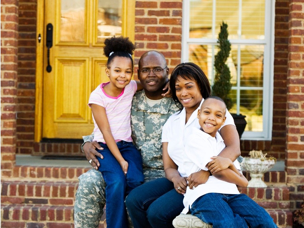A happy military family of four posing in front of a brick house, symbolizing stability and the transition to civilian life.